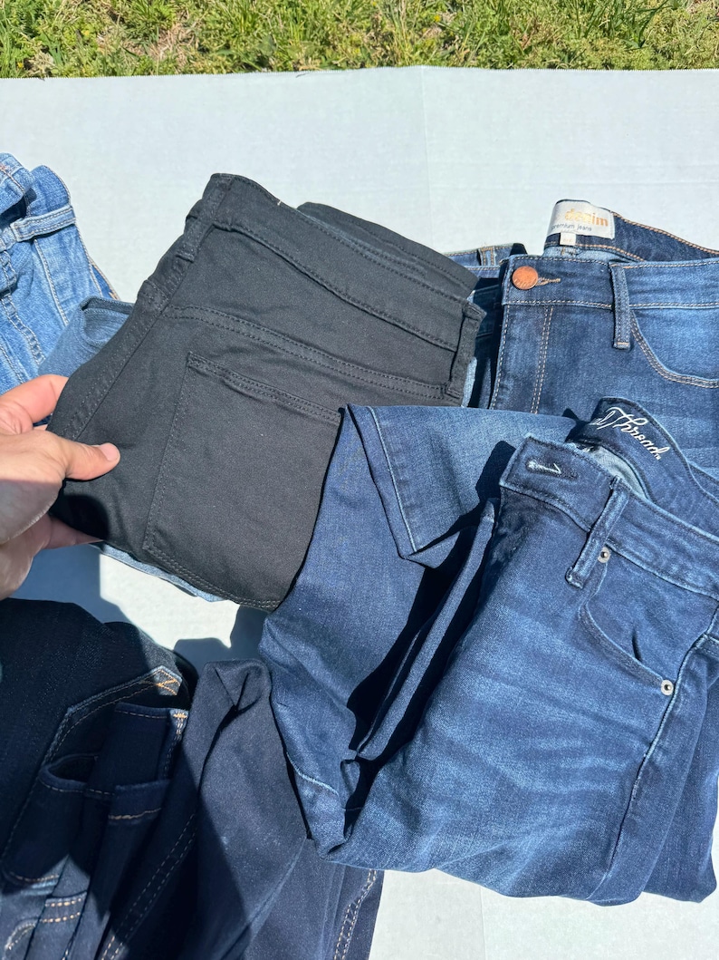 May include: A collection of denim jeans in various shades of blue and black. The jeans are folded and stacked, showcasing different styles and washes. The image is taken outdoors, with a glimpse of green grass in the background.