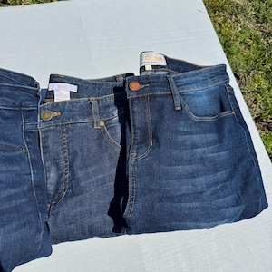 May include: Three pairs of dark wash denim jeans are displayed. The jeans have classic five-pocket styling, with a button and zipper closure. The denim has a slight fade, and the stitching is a contrasting color. The jeans are folded neatly.
