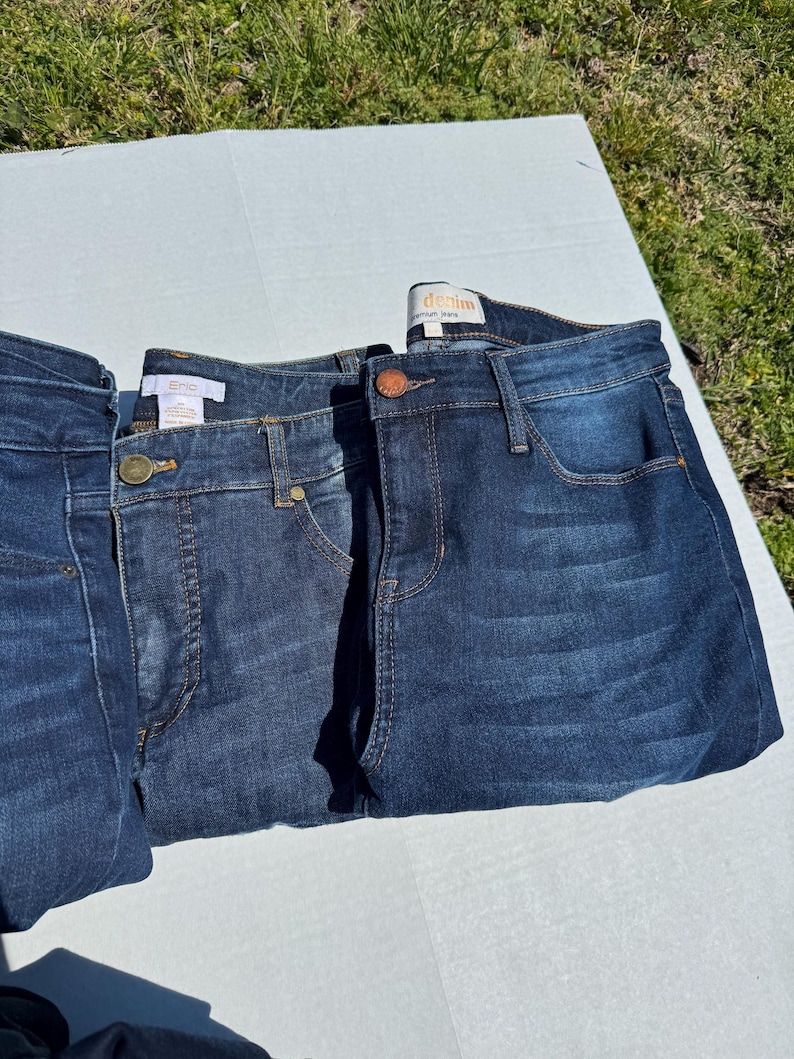 May include: Three pairs of dark wash denim jeans are displayed. The jeans have classic five-pocket styling, with a button and zipper closure. The denim has a slight fade, and the stitching is a contrasting color. The jeans are folded neatly.