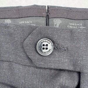 Może przedstawiać: Zbliżenie ciemnoszarych spodni z zapięciem na guzik. Guzik jest srebrny z napisem "VERSACE COLLECTION". Pas ma logo i tekst "VERSACE".
