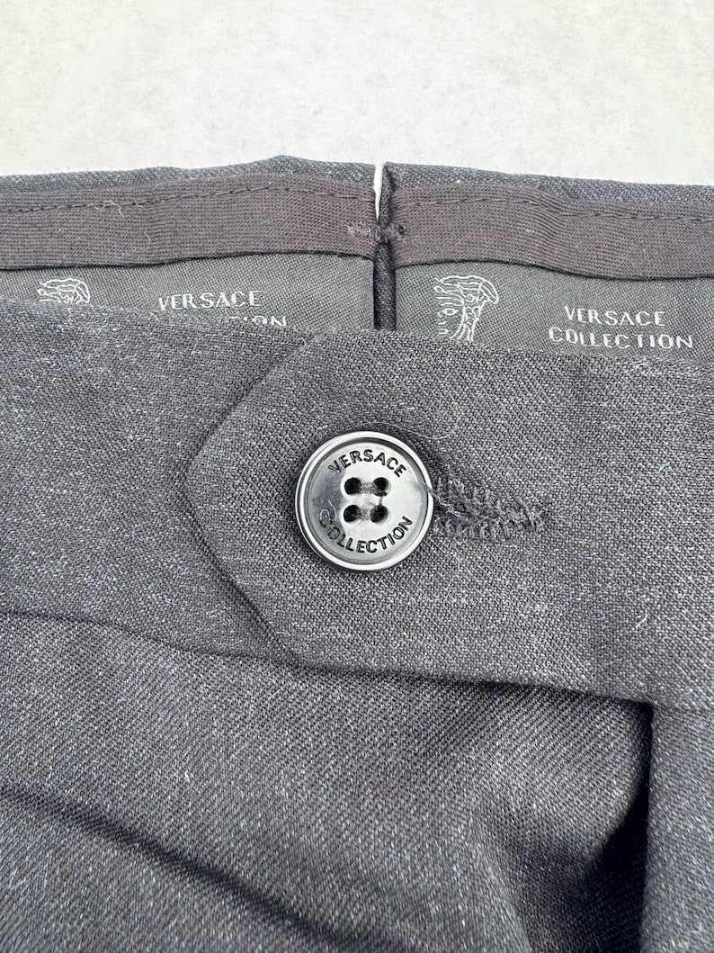 Może przedstawiać: Zbliżenie ciemnoszarych spodni z zapięciem na guzik. Guzik jest srebrny z napisem "VERSACE COLLECTION". Pas ma logo i tekst "VERSACE".