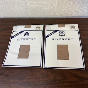 May include: Two Givenchy sheer tights packages with Lycra. The white packaging has a blue border and the Givenchy logo. A rectangular swatch of the tights is visible. The background is a dark brown wooden surface.