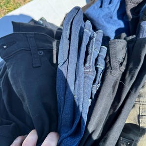 May include: A pile of various denim jeans in shades of blue, black, and olive green. The jeans are folded and stacked, with a hand partially visible, suggesting they are being sorted or displayed. The image is taken outdoors in natural light.
