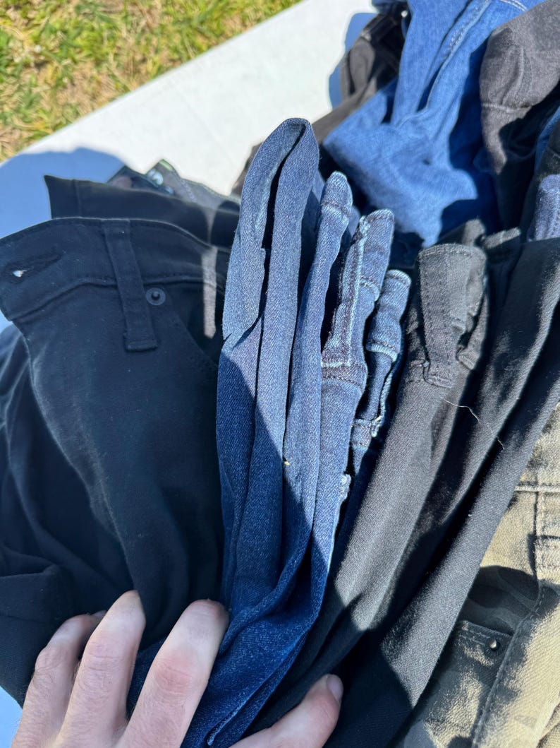 May include: A pile of various denim jeans in shades of blue, black, and olive green. The jeans are folded and stacked, with a hand partially visible, suggesting they are being sorted or displayed. The image is taken outdoors in natural light.