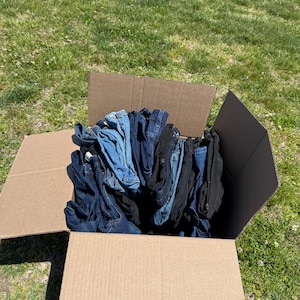May include: A cardboard box filled with folded denim jeans in various shades of blue and black. The box is open, revealing the contents, and sits on a grassy surface under daylight.
