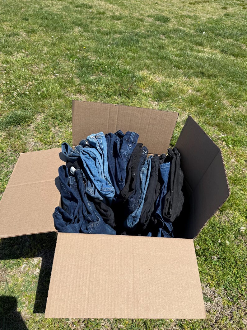 May include: A cardboard box filled with folded denim jeans in various shades of blue and black. The box is open, revealing the contents, and sits on a grassy surface under daylight.