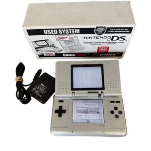 Könnte beinhalten: Ein gebrauchtes silbernes Nintendo DS Handheld-System mit einer GameStop-Box. Auf der Box steht "USED SYSTEM" und "NINTENDO DS". Inklusive Netzteil und Stift. Der Bildschirm zeigt ein Menü an.