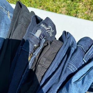 May include: A pile of folded denim jeans in various shades of blue and black. The jeans are stacked together, with the waistbands and pockets visible. One pair has a tag that reads "Universal Thread".