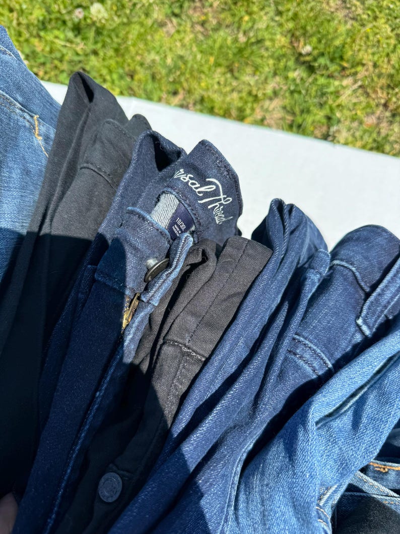 May include: A pile of folded denim jeans in various shades of blue and black. The jeans are stacked together, with the waistbands and pockets visible. One pair has a tag that reads "Universal Thread".