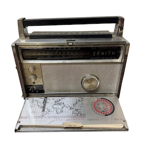 May include: Vintage Zenith Trans-Oceanic radio with a silver and black exterior. The radio features a handle, dials, and a tuning dial. A fold-out map and chart panel is visible. The radio is a classic piece of electronics.