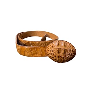 May include: A tan leather belt with a textured crocodile pattern. The belt has a large, oval buckle with a similar crocodile texture. The belt is coiled, showing the full length and design.