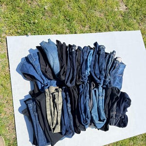 May include: A collection of folded denim jeans in various shades of blue, black, and a camouflage pattern. The jeans are arranged on a white surface, with a grassy background.