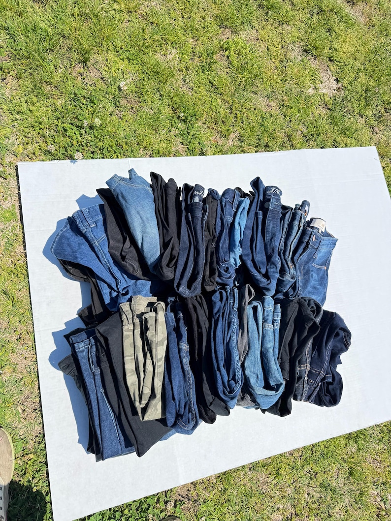 May include: A collection of folded denim jeans in various shades of blue, black, and a camouflage pattern. The jeans are arranged on a white surface, with a grassy background.