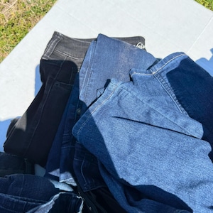 May include: A pile of denim jeans in various shades of blue and black. The jeans are folded and stacked, with visible pockets, seams, and waistbands. The image is taken outdoors, with a white surface and green grass in the background.