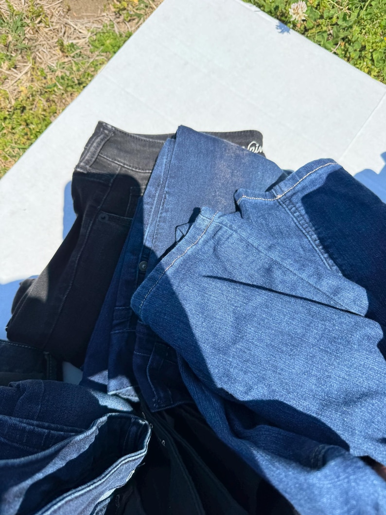 May include: A pile of denim jeans in various shades of blue and black. The jeans are folded and stacked, with visible pockets, seams, and waistbands. The image is taken outdoors, with a white surface and green grass in the background.