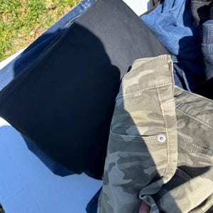 May include: A pile of folded pants in various colors. Included are black, blue, and camouflage-patterned pants. The camouflage pants have a buttoned pocket. The pants are on a white surface.
