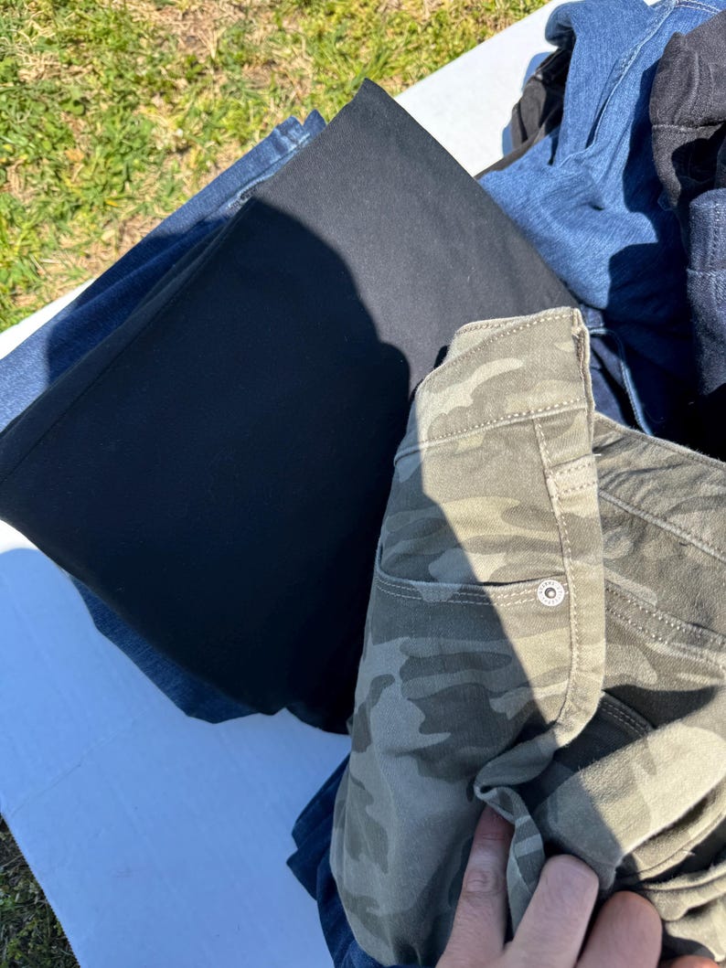 May include: A pile of folded pants in various colors. Included are black, blue, and camouflage-patterned pants. The camouflage pants have a buttoned pocket. The pants are on a white surface.