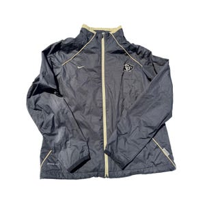 May include: Black Nike Storm-Fit jacket with a full-length zipper and a stand-up collar. The jacket features a gold trim along the zipper, collar, and cuffs. A logo is on the left chest. The jacket is designed for weather protection.