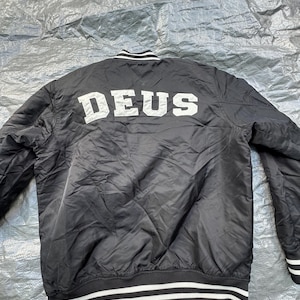 Puede incluir: Chaqueta bomber negra con rayas blancas en el cuello, pu&ntilde;os y dobladillo. La palabra "DEUS" est&aacute; impresa en letras blancas grandes en la espalda. La chaqueta es de un material brillante.