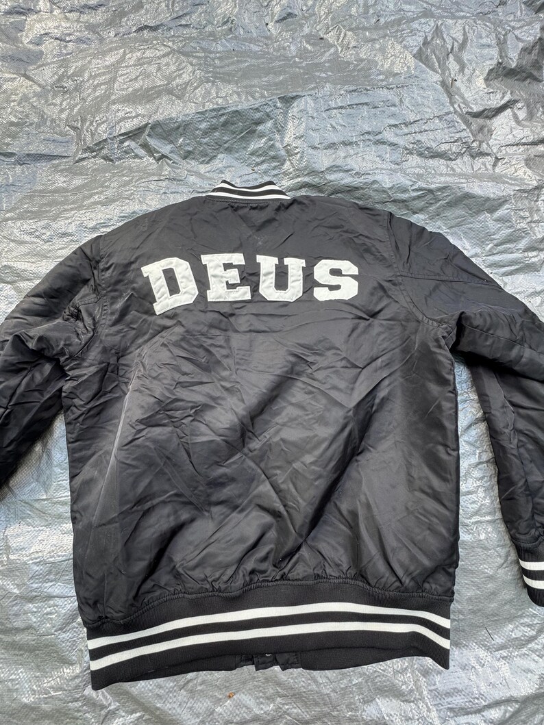 Puede incluir: Chaqueta bomber negra con rayas blancas en el cuello, pu&ntilde;os y dobladillo. La palabra "DEUS" est&aacute; impresa en letras blancas grandes en la espalda. La chaqueta es de un material brillante.