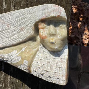 May include: A carved stone sculpture of a person with long hair and a detailed face. The sculpture has a light beige color with red and white accents. The person's clothing is also detailed with carved patterns.