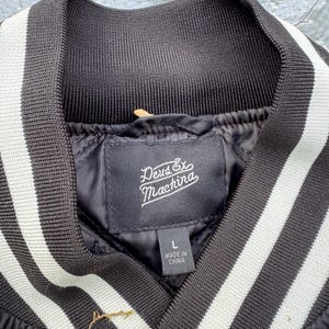 Puede incluir: Chaqueta bomber negra con cuello acanalado y rayas blancas. La chaqueta presenta una etiqueta con el texto "Deus Ex Machina" y "Made in China". La chaqueta es talla L.