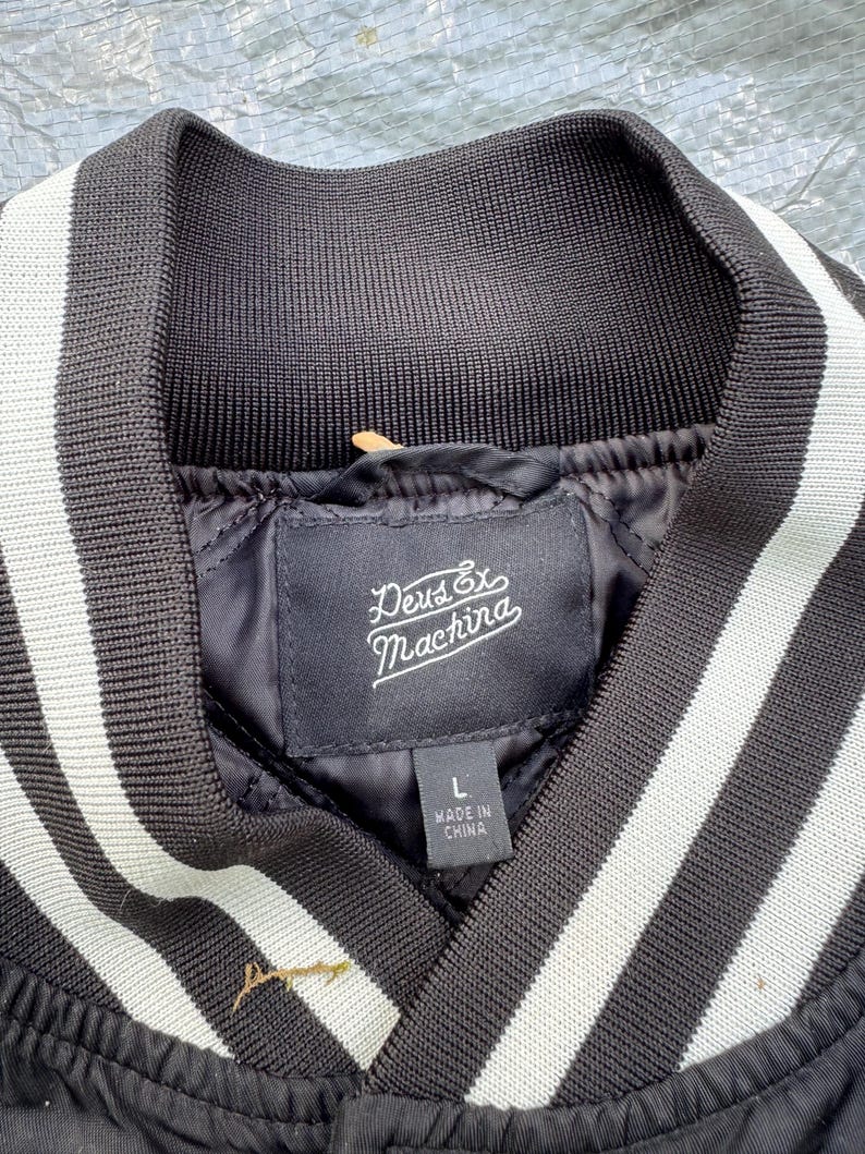 Puede incluir: Chaqueta bomber negra con cuello acanalado y rayas blancas. La chaqueta presenta una etiqueta con el texto "Deus Ex Machina" y "Made in China". La chaqueta es talla L.