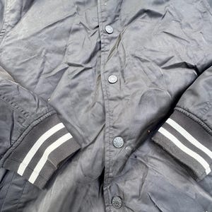 Puede incluir: Chaqueta bomber negra con rayas blancas en los pu&ntilde;os y el cuello. La chaqueta tiene una botonadura frontal y un dobladillo acanalado. Las mangas son largas y la tela parece ser ligera.