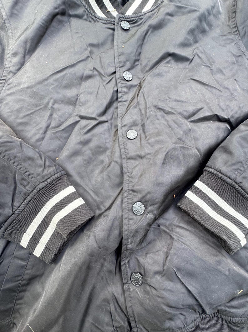 Puede incluir: Chaqueta bomber negra con rayas blancas en los pu&ntilde;os y el cuello. La chaqueta tiene una botonadura frontal y un dobladillo acanalado. Las mangas son largas y la tela parece ser ligera.