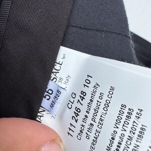 Może przedstawiać: Zbliżenie ciemnoszarego ubrania z białą metką. Metka zawiera tekst, w tym "VERSACE.CERTILOGO.COM" i kod produktu. Widoczna jest tkanina ubrania, o subtelnej fakturze. Etykieta zawiera r&oacute;wnież tekst "CLG 111 246 748 101".
