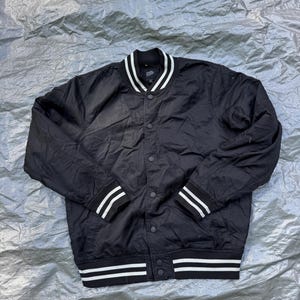Puede incluir: Chaqueta bomber negra con rayas blancas en el cuello, los pu&ntilde;os y el dobladillo. La chaqueta tiene una parte delantera con botones y un estilo universitario cl&aacute;sico. La chaqueta est&aacute; hecha de un material liso, posiblemente nailon.