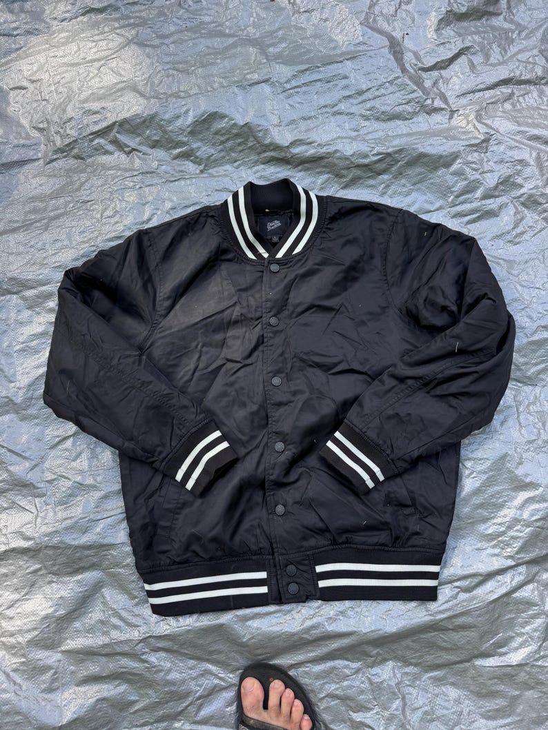 Puede incluir: Chaqueta bomber negra con rayas blancas en el cuello, los pu&ntilde;os y el dobladillo. La chaqueta tiene una parte delantera con botones y un estilo universitario cl&aacute;sico. La chaqueta est&aacute; hecha de un material liso, posiblemente nailon.