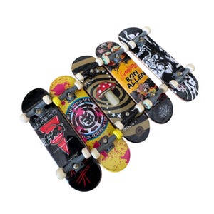 May include: A collection of five miniature skateboards, each with unique designs. The boards feature various graphics, including a mushroom, abstract patterns, and text. They have black trucks and white wheels.