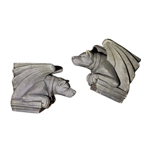 May include: Two gray gargoyle sculptures, each with a dog-like head, wings, and a collar. The gargoyles are in a resting position, with their paws resting on a ledge. The sculptures are made of a stone-like material.