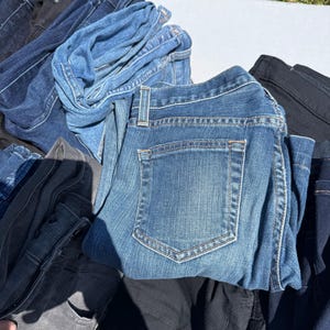 May include: A pile of various denim jeans in shades of blue, black, and gray. The jeans are folded and stacked, showcasing the different washes and styles. The image is taken outdoors in natural light.