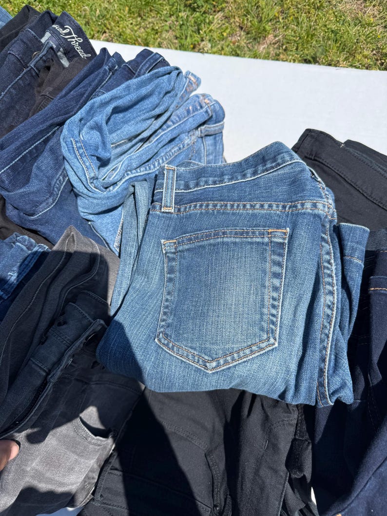 May include: A pile of various denim jeans in shades of blue, black, and gray. The jeans are folded and stacked, showcasing the different washes and styles. The image is taken outdoors in natural light.