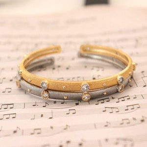 May include: Two gold and silver bracelets with small clear stones. The bracelets are stacked on top of each other and are on a sheet of music.