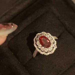 May include: A red gemstone ring with a double halo of smaller white gemstones set in a white metal band.