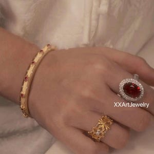 May include: A hand adorned with gold jewelry, including a bracelet with red gemstones, a floral ring, and a large oval-cut red gemstone ring with a halo of smaller stones. The jewelry is set against a soft, neutral background.