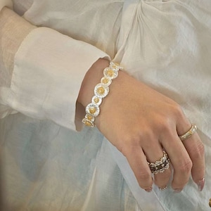 May include: A silver and gold bracelet featuring a daisy design. The bracelet is worn on a wrist, with a white long-sleeved shirt in the background. Several rings are also visible on the hand, including a gold band and a multi-stone ring.
