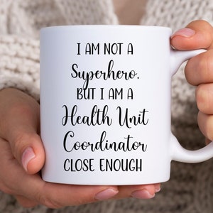 May include: White ceramic mug with black text that reads "I am not a superhero. But I am a Health Unit Coordinator close enough."