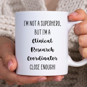 May include: White ceramic mug with black text that reads "I'm not a superhero, but I'm a Clinical Research Coordinator close enough!"