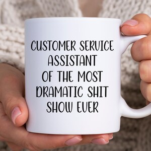 May include: White ceramic mug with black text that reads "Customer Service Assistant of the Most Dramatic Shit Show Ever".