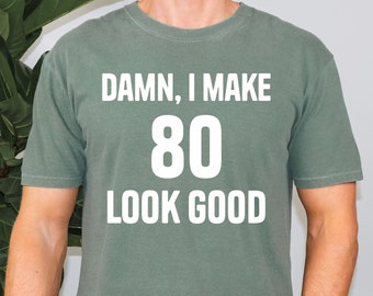 Puede incluir: Una camiseta verde con el texto "DAMN, I MAKE 80 LOOK GOOD" impreso en blanco.