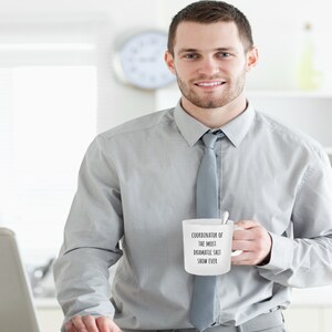 May include: White ceramic coffee mug with black text that reads "Coordinator of the most dramatic shit show ever". A man in a light gray button-down shirt and a gray tie is holding the mug.