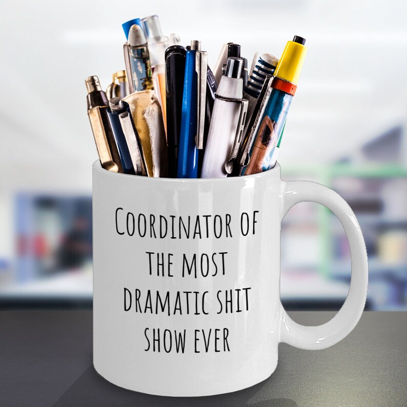 May include: White ceramic mug with black text that reads "Coordinator of the most dramatic shit show ever". The mug is filled with pens and pencils.