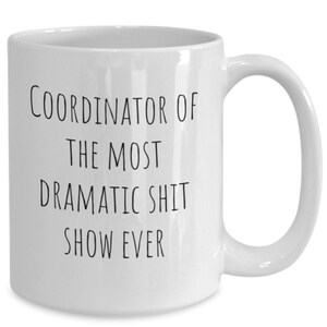 May include: White ceramic coffee mug with black text that reads "Coordinator of the most dramatic shit show ever".