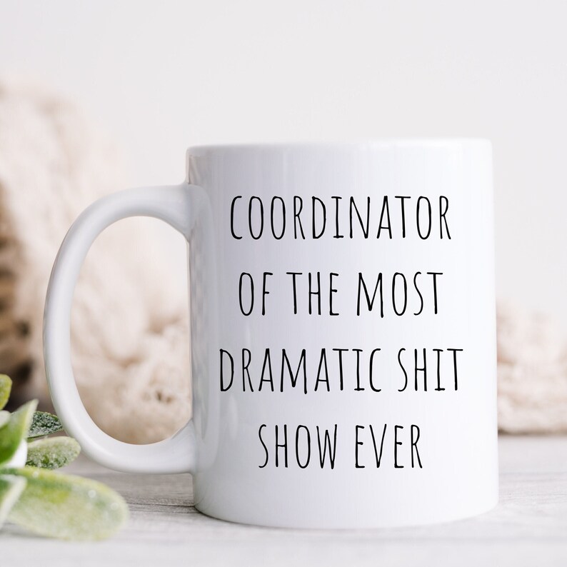 May include: White ceramic coffee mug with black text that reads "Coordinator of the most dramatic shit show ever".