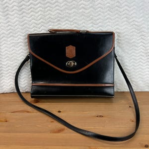 Puede incluir: Maletín de cuero negro con ribete marrón y bandolera. El bolso presenta un emblema marrón en la solapa delantera y un cierre plateado. El asa es marrón. El bolso está sobre una superficie de madera.