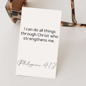 May include: A white rectangular card featuring the black printed text I can do all things through Christ who strengthens me and Philippians 4:13, positioned partially behind a pair of brown and black tortoiseshell patterned sunglasses.